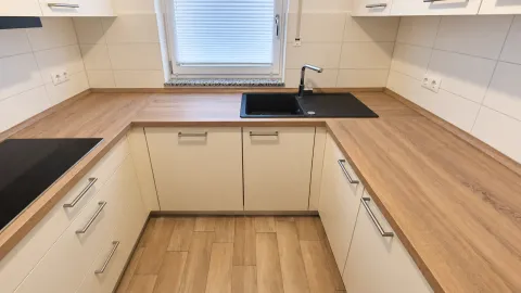 Weiße U-förmige Kundenküche mit Holzoptik-Arbeitsplatte, Kochfeld links und schwarzer Spüle am Fenster